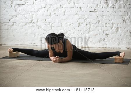 Young yogi attractive woman practicing yoga concept, doing advanced Straight Angle exercise, Samakonasana pose with Seated Forward Bend using blocks, working out, full length, white loft background