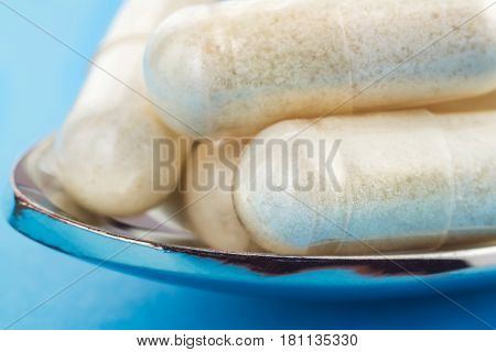 White capsules of glucosamine chondroitin healthy supplement pills in the spoon on blue background macro image