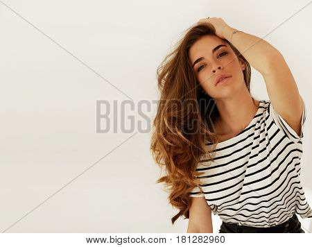 Attractive Caucasian female student in sailor shirt joyfully tying her long dark hair with mouth wide open looking with serious expression at the camera posing against white wall. Copy space for text