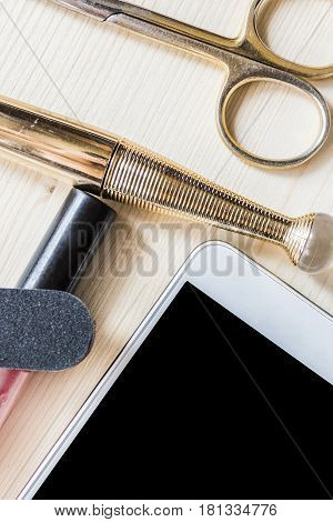 White mobile phone, mascara in a golden tube, gold manicure scissors, pink lip gloss and a nail file on a light wooden table. Women's tricks concept