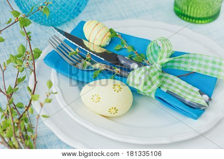 Easter table setting with painted eggs green spring branch and cutlery on light blue wooden background. Spring table setting. Easter dinner decoration. Happy easter holiday decoration concept.
