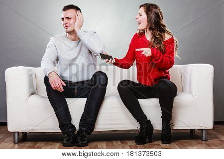 Angry furious wife shouting at husband showing text messages from lover mistress on his mobile phone. Outraged girlfriend find out about boyfriend affair romance betrayal. Cheating man covering ears.