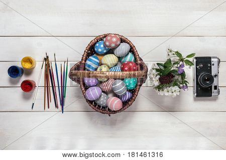Basket with colorful Easter eggs, painted in handmade, bouquet of spring flowers, paints, brushes and retro camera stand in a row on an old white wooden background. Top view, flat lay style