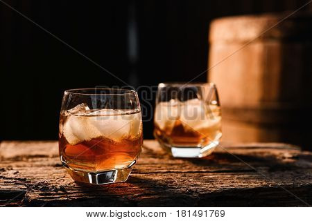 A glass of whiskey with ice on a wooden background close-up in the background of a barrel of whiskey
