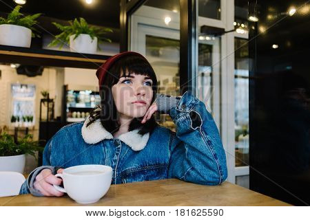 Stylish young hipster girl sitting in a cafe drinking coffee and looking in the window and dreaming