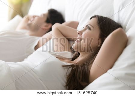 Side view of happy woman looking up lying in comfortable bed with beloved man, cute married couple in white shirts resting on bed with hands behind the head after waking up in the morning