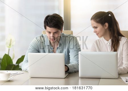 Young lady taking inquisitive glance, sneak peek at laptop screen of male colleague, stealing private information from computer of competitor, nosy female coworker sitting next to man spying on him