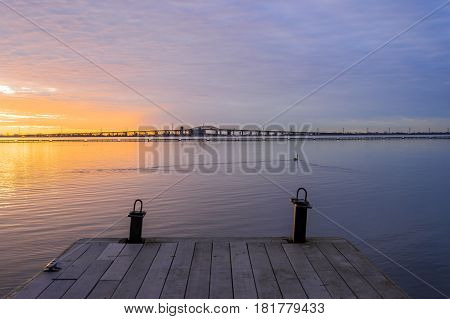 Wooden boat pier leading out to vibrant colorful sunrise over tranquil waters bridge at horizon