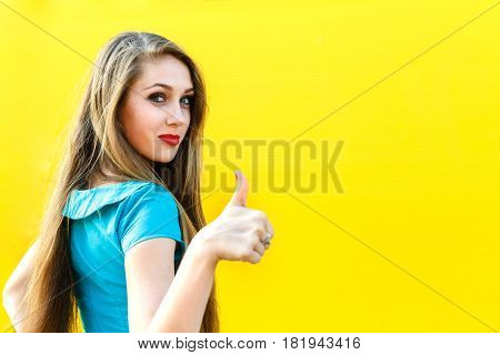 beautiful blonde girl in blue dress showing finger up sign OK on a yellow background.