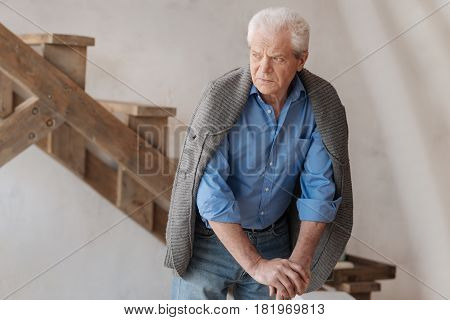 Thoughtful look. Unhappy grey haired elderly man standing in the room and leaning on the walking stick while looking aside