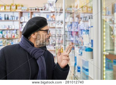 Old Man In Glasses Looking At Pharmacy Shelf With Medical Products And Pointing With Fingers. Custom