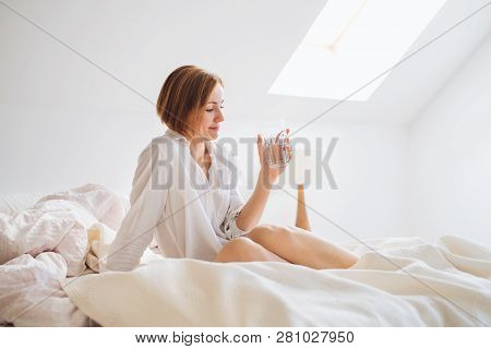 A Young Woman With Night Shirt Sitting Indoors On Bed In The Morning, Holding Water.