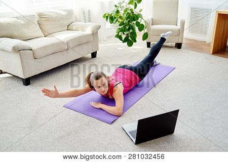Beautiful Young Woman Doing Exercise For Back On Floor At Home, Online Training On Laptop Computer, 