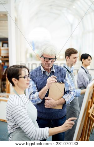 Portrait Of Mature Art Teacher Helping Group Of Students Painting Picture On Easel In Art Class