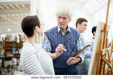 Portrait Of Mature Art Teacher Frowning While Helping Student Painting Picture On Easel In Art Class