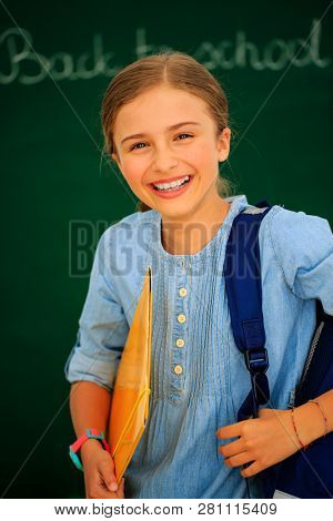 Back to school.. Funny beatifull girl sitting in the clasroom against blacboard. Child in school with book and accessories. Education concept