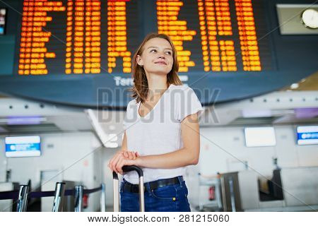 Young Woman In International Airport With Luggage Near Flight Information Display