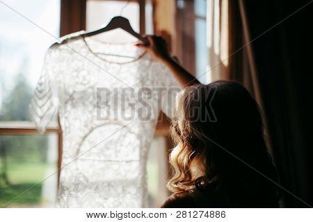 Blonde Woman With Curly Hair Holding And Looking At Beautiful White Wedding Dress On A Hanger, Stand