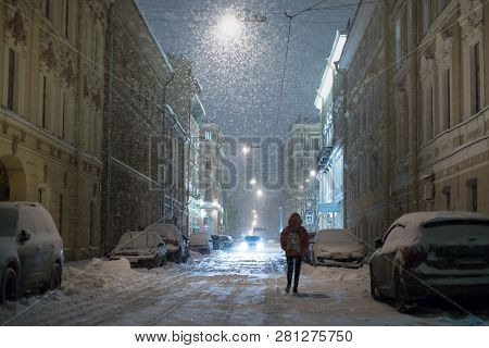 Silhouette Of Female Walking Along Parked Cars On Empty Evening City Street During A Snowstorm, Sele