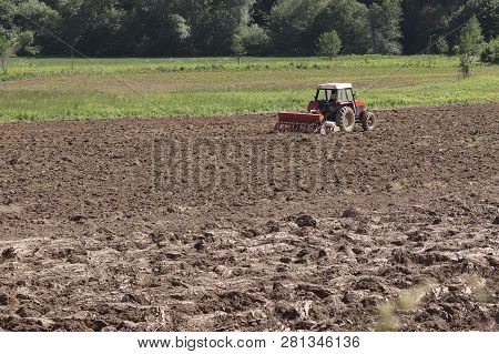 A Farmer On A Red Tractor With A Seeder Sows Grain In Plowed Land In A Private Field In The Village 