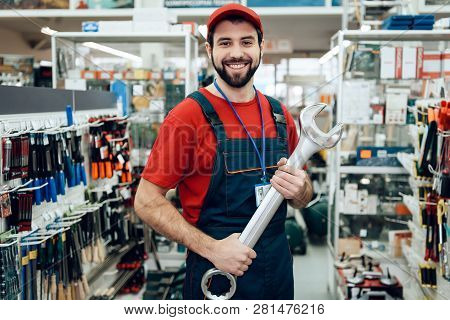 Salesman In Red Shirt And Baseball Cap Is Posing With New Giant Wrench In Power Tools Store.