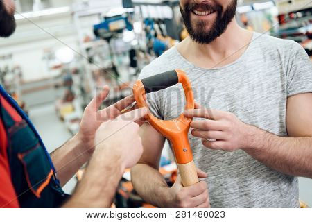 Salesman In Red Shirt And Baseball Cap Is Showing Couple Of Clients New Showel In Power Tools Store.