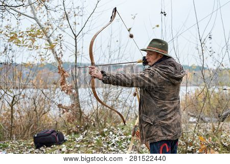 Man In The Hat Shoots From Wooden Bow On Outside, In Autumn