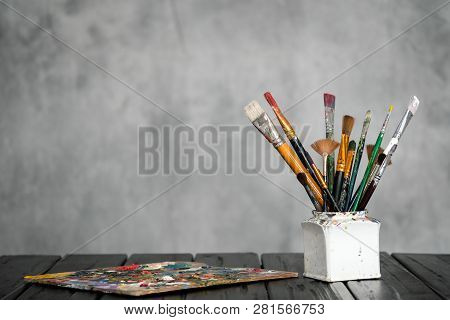 Artists Tools, Brushes, Paints And A Palette Lie On A Black Wooden Table On A Gray Fabric Background