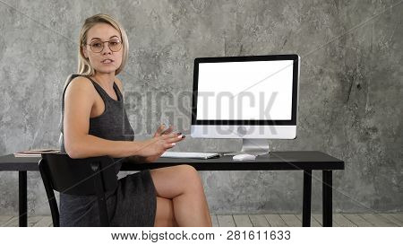 Cheerful Young Woman Sitting In Office Talking To Camera And Showing On Display Of Computer Somethin