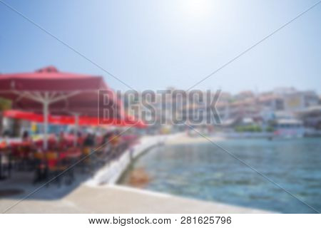 Restaurant Tables And Chairs Below Red Sunshades On The Terrace On The Seashore. Blurred Scene Of Th