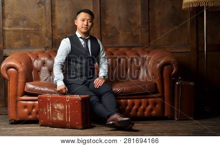 A Young Man In A Suit Sits On A Leather Brown Sofa With A Suitcase