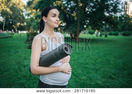 Cheeerful Nice Young Oregnant Woman Stand In Green Park. She Hold Rolled Yoga Mate And Keep Hands On