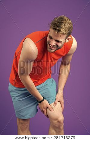 Sportsman With Knee Pain On Violet Background. Man In Orange Vest And Blue Shorts. Health Problem An