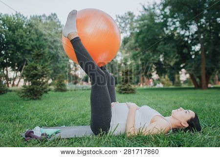 Concentrated Young Pregnant Woman Exerising On Yoga Mate In Park. She Hold Big Orange Fitness Ball B