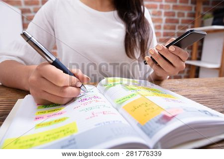 Close-up Of A Businesswoman Holding Cellphone Writing Schedule In The Diary With Pen