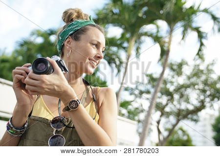 Stylish young woman taking photos with vintage camera surrounded by palm trees