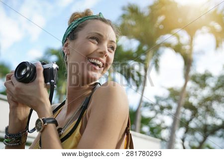 Stylish young woman taking photos with vintage camera surrounded by palm trees