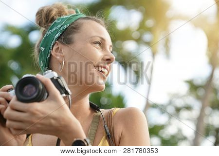 Stylish young woman taking photos with vintage camera surrounded by palm trees