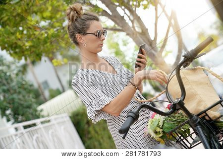 Pretty young woman texting next to bicycle outdoors in leafy city