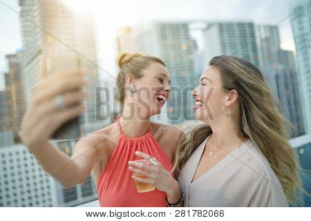 Two attractive funloving friends taking selfie on night out at roofttop bar in city