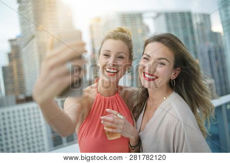 Two attractive funloving friends taking selfie on night out at roofttop bar in city