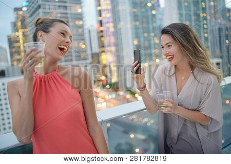 Smiling young woman taking photo of funloving friend on rooftop bar in city