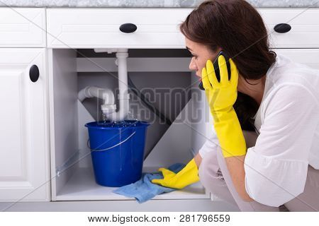 Sad Young Woman Calling Plumber In Front Of Water Leaking From Sink Pipe