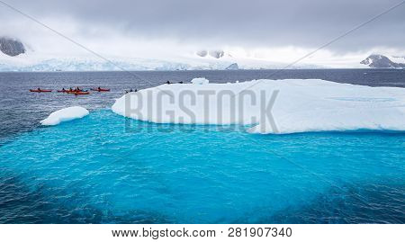 Huge Iceberg With Blue Underwater Part And Small Tourists Kayaks With Antarctic Lagoon In The Backgo
