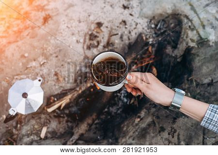 Girls Hand Holds A Hot Cup Of Coffee At The Background Of The Campfire. Concept Adventure Active Vac