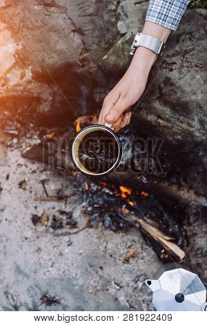 Girls Hand Holds A Hot Cup Of Coffee At The Background Of The Campfire. Concept Adventure Active Vac