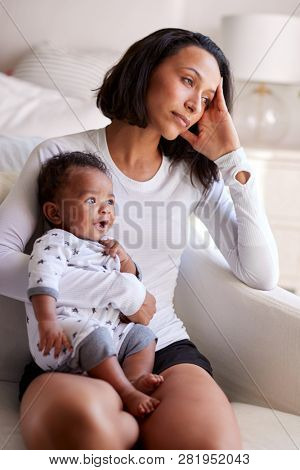 Young adult mother sitting in an armchair in her bedroom, holding her three month old baby son, looking away in contemplation, vertical