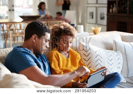 Pre-teen girl sitting on sofa in the living room using tablet computer with her father, mother and toddler sitting at a table in the background, selective focus