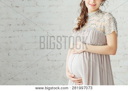 Portrait Of A Elegant Unrecognizable Pregnant Woman Wearing Grey Dress And Posing In White Studio. P