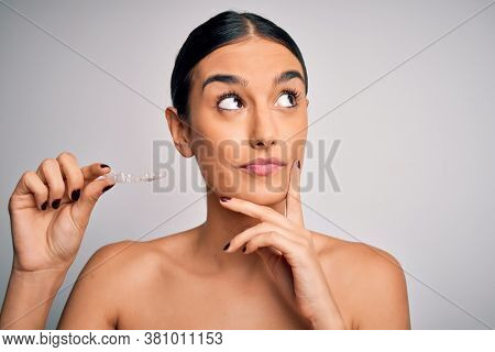 Young beautiful brunette woman holding dental aligner orthodontic to teeth corretion serious face thinking about question, very confused idea